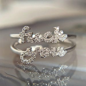Puede incluir: Dos anillos de plata apilados. El anillo superior deletrea la palabra "fuck" y el anillo inferior la palabra "you", ambos en escritura cursiva. Ambos anillos están incrustados con pequeñas gemas brillantes.