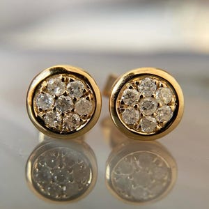 May include: A pair of gold stud earrings. Each earring features a cluster of small, clear gemstones set within a circular gold frame. The earrings are displayed on a reflective surface.