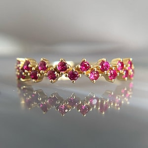 Könnte beinhalten: Ein goldener Ring mit einem Zickzackmuster aus kleinen, runden, leuchtend rosa Edelsteinen. Die Reflexion des Rings ist darunter sichtbar. Der Hintergrund ist verschwommen und neutral. Ein Schmuckstück.
