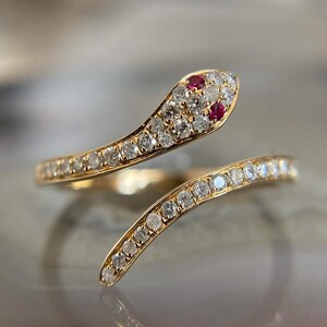 Può includere: Un anello a forma di serpente dorato, con testa e corpo incastonati di piccoli diamanti scintillanti. La testa del serpente presenta due gemme rosse e l'anello si avvolge attorno al dito. Un primo piano mette in risalto l'intricato design e la maestria dell'anello.