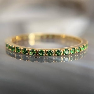 May include: A gold band ring with a continuous row of small, vibrant green gemstones. The ring is set against a blurred background, highlighting the ring's design and the sparkle of the gemstones. The ring is a piece of jewelry.