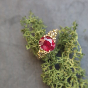 May include: A gold ring with a large, red ruby gemstone and smaller diamonds on the band. The ring is set on a bed of green moss.