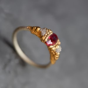 May include: A gold ring with a red gemstone and two smaller white gemstones. The ring has a textured band.