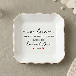 Può includere: Un vassoio portagioie in ceramica bianca con bordo dorato, con la scritta "we love" e un versetto biblico. Sono presenti anche i nomi Sophia & Ethan e l'anno 2026. Elementi decorativi: fiori e perle.