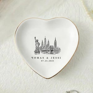 May include: A heart-shaped, white ceramic trinket dish with a gold rim. The dish features a black and white illustration of the New York City skyline, including the Statue of Liberty and the Brooklyn Bridge. The names "Thomas & Jessi" and the date "07.25.2025" are printed below the skyline.