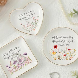 Puede incluir: Tres platos de cerámica blancos para anillos en forma de corazón, cuadrado y redondo, cada uno con borde dorado y diseño floral. El texto "A Sweet Friendship Refreshes The Soul Proverbs 27:9" está impreso.