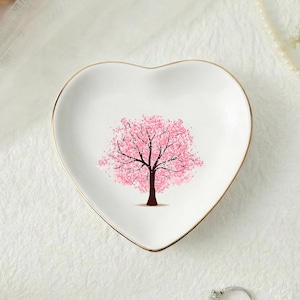 以下が含まれることがあります： 金色の縁取りが付いた、ハート型の白いセラミック製の小物入れ。中央にはピンク色の桜の木の模様があります。花やジュエリーなど、装飾的な要素に囲まれています。