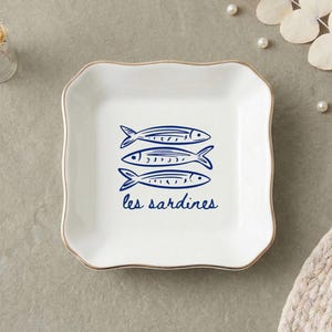 May include: White ceramic dish with a scalloped edge and gold trim. The dish features three blue fish illustrations and the text "les sardines". The dish is square and sits on a neutral surface.