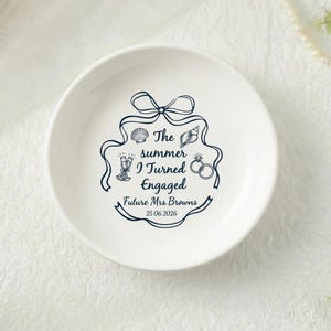 Może przedstawiać: Biała ceramiczna miseczka na pierścionki z granatowym wzorem. Miseczka zawiera napis "The summer I turned engaged" z ilustracjami muszli, kieliszków szampana i pierścionków. Data "25.06.2026" jest również uwzględniona.