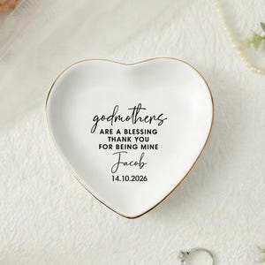 May include: A heart-shaped white ceramic trinket dish with a gold rim. The dish has the inscription "godmothers are a blessing thank you for being mine Jacob 14.10.2026" in black script. The background includes flowers and jewelry.