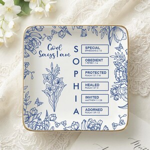 Puede incluir: Un plato cuadrado de cerámica blanca con borde dorado, decorado con ilustraciones florales azules y el texto God Says I am SOPHIA, junto a las palabras SPECIAL, OBEDIENT, PROTECTED, HEALED, INVITED y ADORNED con sus versículos bíblicos.