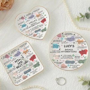 Könnte beinhalten: Drei Keramik-Schmuckschalen mit goldenen Rändern, jede mit Affirmationen. Eine ist herzförmig, eine quadratisch und eine rund. Die Schalen sind weiß mit farbigem Text und Designs. Ein silberner Ring und Blumen sind ebenfalls im Bild.