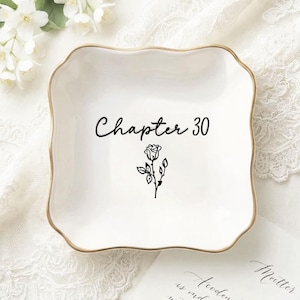 May include: A square white ceramic trinket dish with scalloped edges and a gold-toned rim. The center of the dish features the black text "Chapter 30" written in a cursive script, positioned directly above a small, simple black line drawing of a rose.