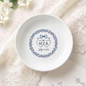 Peut inclure: Petit plat rond en céramique blanche avec un motif de couronne florale bleue. Il présente deux cœurs connectés avec les lettres "M" et "R" et la date "16 juillet 2026". Un nœud bleu est au-dessus des cœurs.