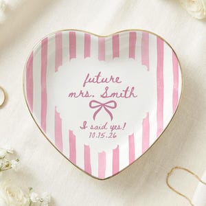 Peut inclure: Un vide-poche en céramique en forme de cœur avec des rayures verticales roses et blanches et un bord doré. Le centre présente le texte future mrs. Smith, un motif de nœud, la phrase I said yes! et la date 10.15.26 en écriture rose.