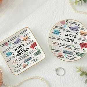 May include: Two decorative ceramic trays with gold trim. One is square and reads "Nova's Daily Reminder" with affirmations. The other is round and says "Lucy's Badass Affirmations". Both have colorful text and designs.