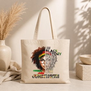 May include: A beige tote bag featuring a graphic of a Black woman with glasses and the words "I AM BLACK HISTORY." Additional text includes "Beautiful, Powerful, Resilient, Magical, Smart, Loved" and "Juneteenth."