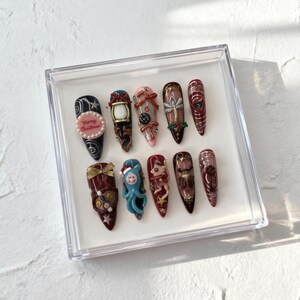 May include: A set of twelve long, almond-shaped artificial nails, each with a unique design. The designs include Christmas-themed elements like a lantern, a gift, and the text "Merry Christmas". The nails are displayed in a clear plastic box.