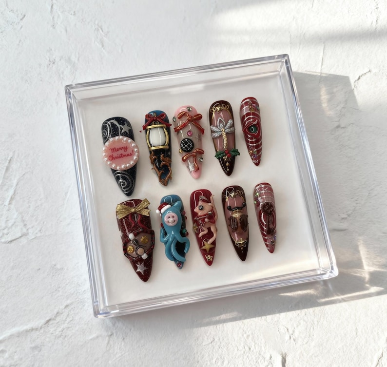 May include: A set of twelve long, almond-shaped artificial nails, each with a unique design. The designs include Christmas-themed elements like a lantern, a gift, and the text "Merry Christmas". The nails are displayed in a clear plastic box.