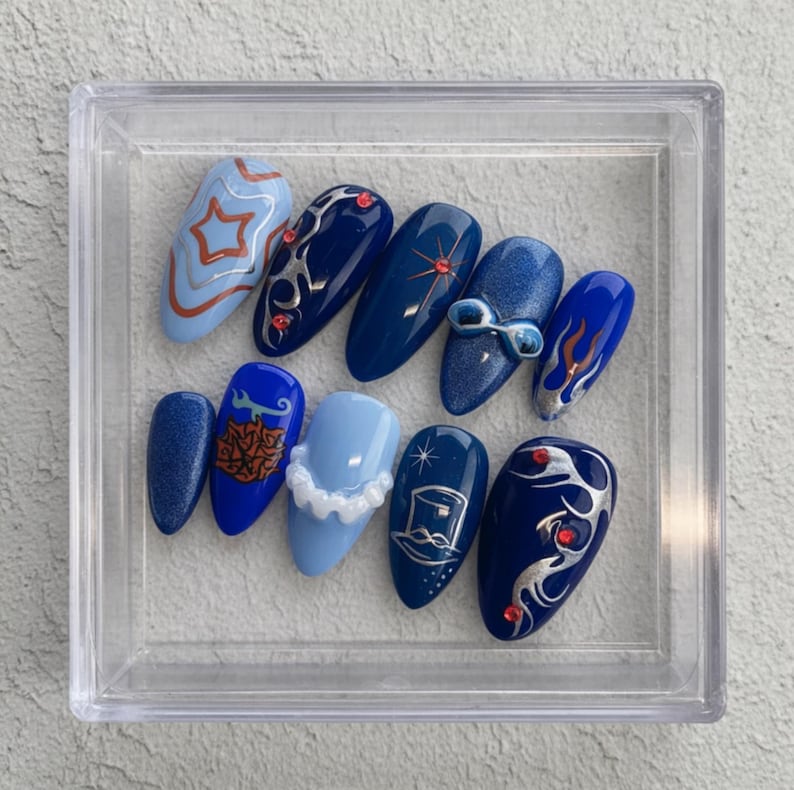 May include: A set of ten artificial nails in a clear plastic case. The nails are various shades of blue, with designs including stars, swirls, and flame-like patterns. Some nails feature silver and red accents.