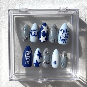 May include: A collection of ten false nails in a clear case. The nails are decorated with designs including blue and white stars, the text "TWICE", "THIS IS FOR", and the number "4". Some nails have silver glitter and wire detailing.