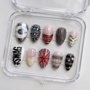 Könnte beinhalten: Ein Set aus zehn künstlichen Nägeln in einer transparenten Kunststoffbox. Die Nägel weisen verschiedene Designs auf, darunter eine Union Jack, metallische Akzente und das Wort "SWAG". Farben sind Silber, Rosa, Blau und Schwarz.