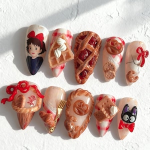 May include: Ten artificial nails with diverse designs. The designs feature a cartoon character, a waffle with cream, a cherry pie, a pretzel, biscuits, and a bow. The nails are painted in various colours, including red, white, and brown, creating a whimsical aesthetic.
