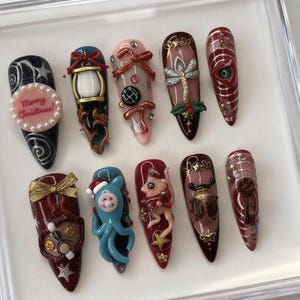 May include: A collection of ten long, decorated fingernails. Designs include a lantern, bows, a dragonfly, and a Christmas message. Colors include red, blue, gold, and pink. Some nails feature 3D embellishments and glitter. The nails are arranged in a clear display case.