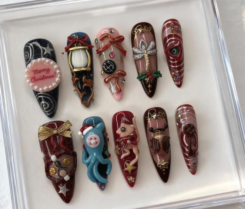 May include: A collection of ten long, decorated fingernails. Designs include a lantern, bows, a dragonfly, and a Christmas message. Colors include red, blue, gold, and pink. Some nails feature 3D embellishments and glitter. The nails are arranged in a clear display case.