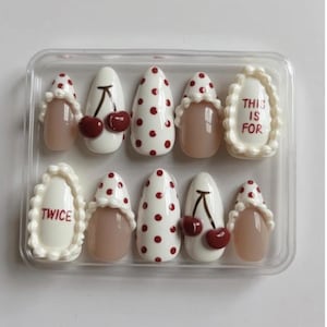 May include: A set of ten press-on nails in a clear plastic case. The nails feature various designs, including white with red polka dots, nude, and white with a cherry design. Some nails have the text "THIS IS FOR" and "TWICE".