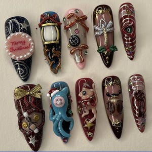 May include: A collection of ten long, almond-shaped acrylic nails with various designs. The nails feature a range of colors, including dark blue, pink, and burgundy, and are adorned with intricate embellishments such as bows, stars, and miniature figurines. One nail has the text "Merry Christmas!"