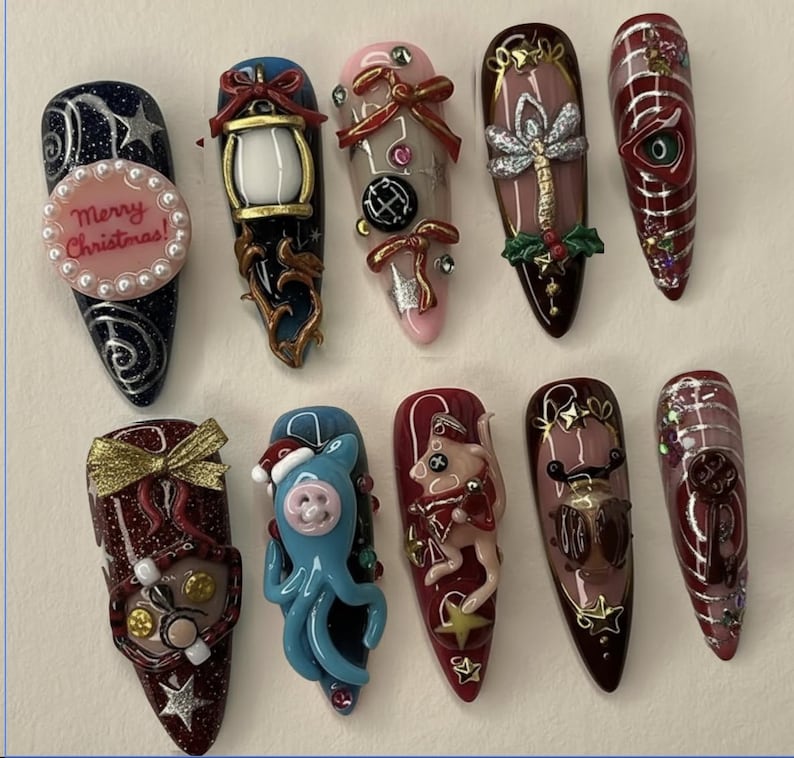 May include: A collection of ten long, almond-shaped acrylic nails with various designs. The nails feature a range of colors, including dark blue, pink, and burgundy, and are adorned with intricate embellishments such as bows, stars, and miniature figurines. One nail has the text "Merry Christmas!"