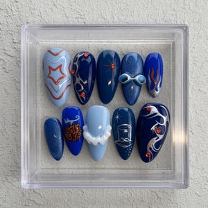May include: A set of ten artificial nails in a clear plastic case. The nails are various shades of blue, with designs including stars, swirls, and a bow. Some nails have glitter or red accents. The nails are oval-shaped.