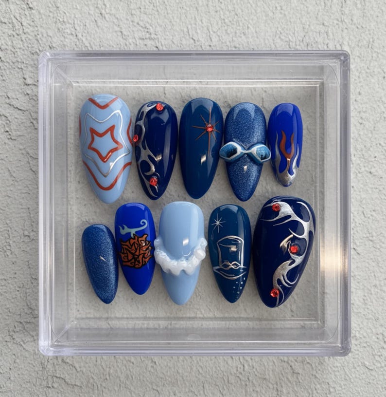 May include: A set of ten artificial nails in a clear plastic case. The nails are various shades of blue, with designs including stars, swirls, and a bow. Some nails have glitter or red accents. The nails are oval-shaped.
