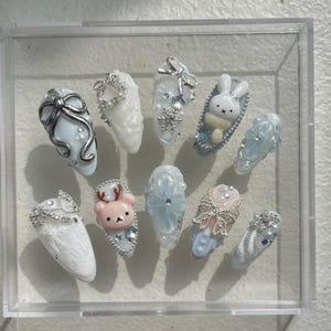 Puede incluir: Un conjunto de diez uñas artificiales con varios diseños, incluyendo lazos, copos de nieve y un conejito. Las uñas son principalmente blancas y celeste, con detalles plateados y adornos. Las uñas se exhiben en una caja de acrílico transparente.