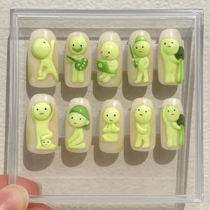 May include: A set of ten press-on nails with a pale yellow base, each featuring a 3D lime green figure in various poses. The figures have simple, cartoonish features and are enclosed in a clear plastic case.