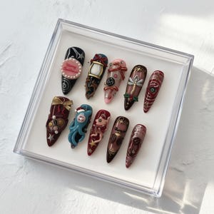 May include: A set of twelve long, almond-shaped artificial nails, each uniquely decorated with intricate designs. The nails feature a variety of colors, including black, red, pink, and gold, with embellishments like bows, pearls, and miniature figurines. The nails are displayed in a clear plastic box.