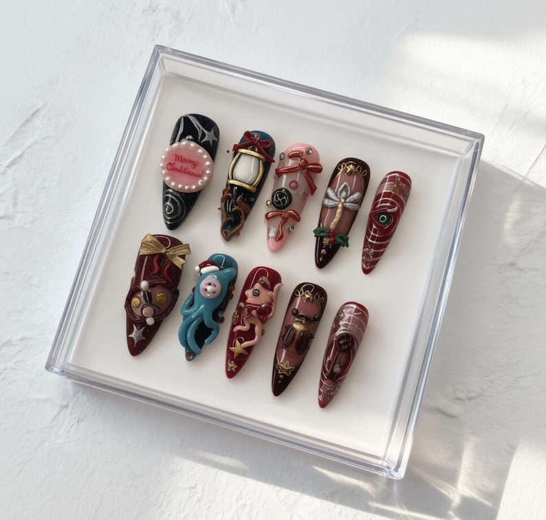May include: A set of twelve long, almond-shaped artificial nails, each uniquely decorated with intricate designs. The nails feature a variety of colors, including black, red, pink, and gold, with embellishments like bows, pearls, and miniature figurines. The nails are displayed in a clear plastic box.