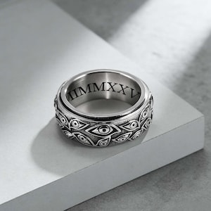 May include: A silver spinner ring with an engraved eye pattern around the band. The inner band features the Roman numeral "IIMMMXXV". The ring is displayed on a white surface.