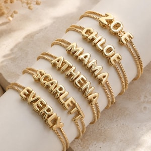 Op de afbeelding: Een collectie van zes goudkleurige kettingarmbanden, elk voorzien van een gepersonaliseerde naam of woord in verhoogde, metalen blokletters. De namen op de armbanden zijn ZOE, CHLOE, MAMA, ATHENA, SCARLETT en EVERLY, verticaal gerangschikt.