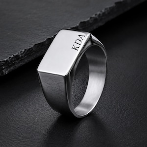 Puede incluir: Un anillo de sello plateado pulido con una cara rectangular plana. La superficie del anillo está grabada con las letras mayúsculas KDA en una fuente serif negra y clara. La banda es lisa y metálica con un acabado reflectante en todo el diseño.