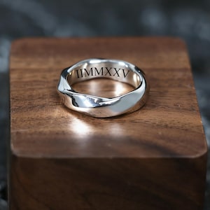 May include: A silver ring with a wavy design. The inside of the ring has the Roman numeral inscription "IIMMMXXV". The ring is displayed on a wooden block.