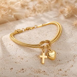 Puede incluir: Una pulsera de cadena de serpiente doble en tono dorado con un nudo central. Del nudo cuelgan dos dijes: una cruz lisa en tono dorado y un dije de corazón liso en tono dorado grabado con el nombre Siomara en una fuente de escritura cursiva.
