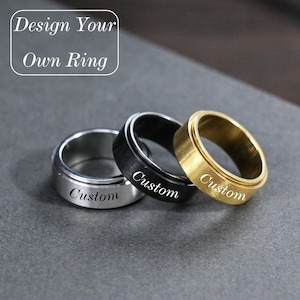May include: Three custom spinner rings in silver, black, and gold. Each ring has the word "Custom" engraved on the band. The image includes the text "Design Your Own Ring" in a white box.