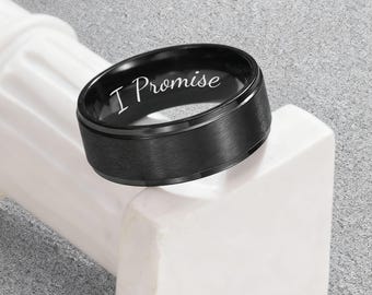 May include: A black ring with the inscription "I Promise" in white lettering. The ring has a brushed metal finish and a simple, modern design. The ring is displayed on a white, geometric surface against a gray background.