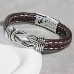 Peut inclure: Bracelet en cuir marron avec un nœud argenté et un fermoir magnétique. Le fermoir est gravé d'une empreinte de main et des mots "To My Amazing Son". Le bracelet a des coutures blanches.