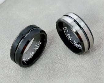 May include: Two black and silver wedding bands. One ring has the inscription "Neil+Mia", the other ring has the inscription "02.06.2025". The rings are on a gray surface.