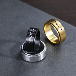 Puede incluir: Tres anillos giratorios personalizados en oro, negro y plata. Cada anillo tiene la palabra "Custom" grabada en la banda exterior. Los anillos se muestran sobre una superficie gris oscuro.