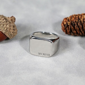 Könnte beinhalten: Ein silberner Siegelring mit quadratischer Oberfläche. Der Ring hat abgerundete Kanten und die römischen Ziffern XV.XI.VII sind auf der Oberfläche eingraviert. Der Ring wird auf einer hellgrauen Oberfläche präsentiert.