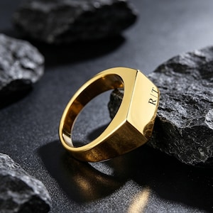 Op de afbeelding: Een gepolijste goudkleurige zegelring met een plat, rechthoekig vlak gegraveerd met de letters R|T. De ring heeft een glad, reflecterend oppervlak en een massief bandontwerp, wat zorgt voor een strakke, metalen afwerking over het hele sieraad.